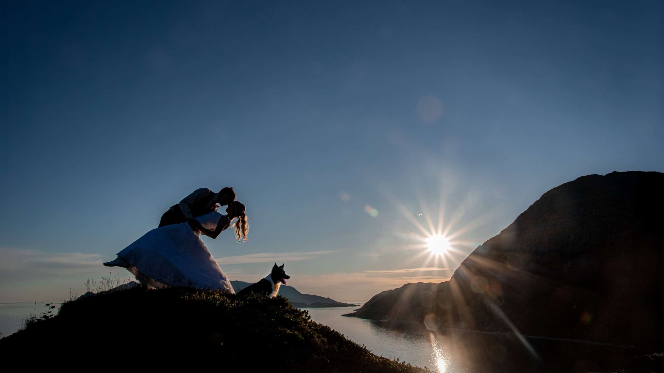 Romantisk bryllupsfoto med brudepar og hund ved solnedgang over fjorden.
