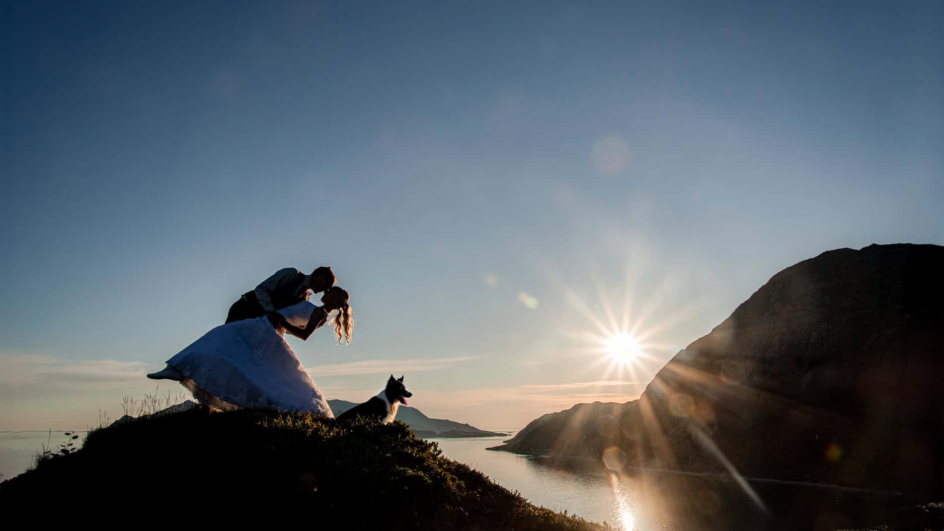 Bryllupsfotografi av et par som kysser på en fjelltopp med utsikt over vannet ved solnedgang.