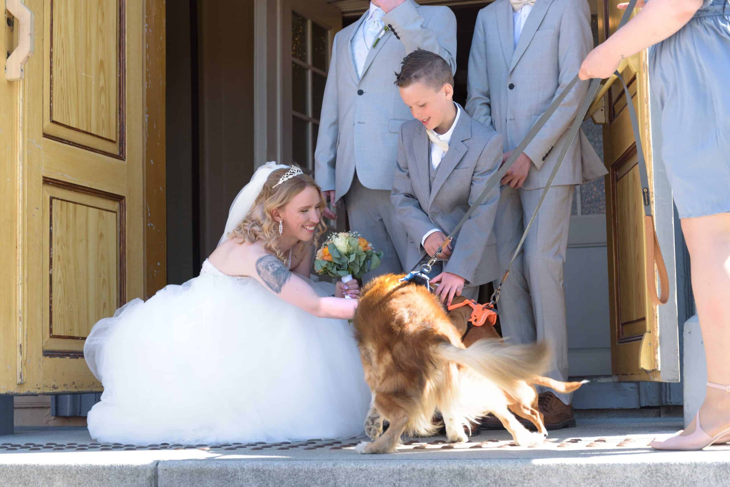 Brud og familie med hund utenfor bryllupslokale, smilende og kledd i formelle klær.